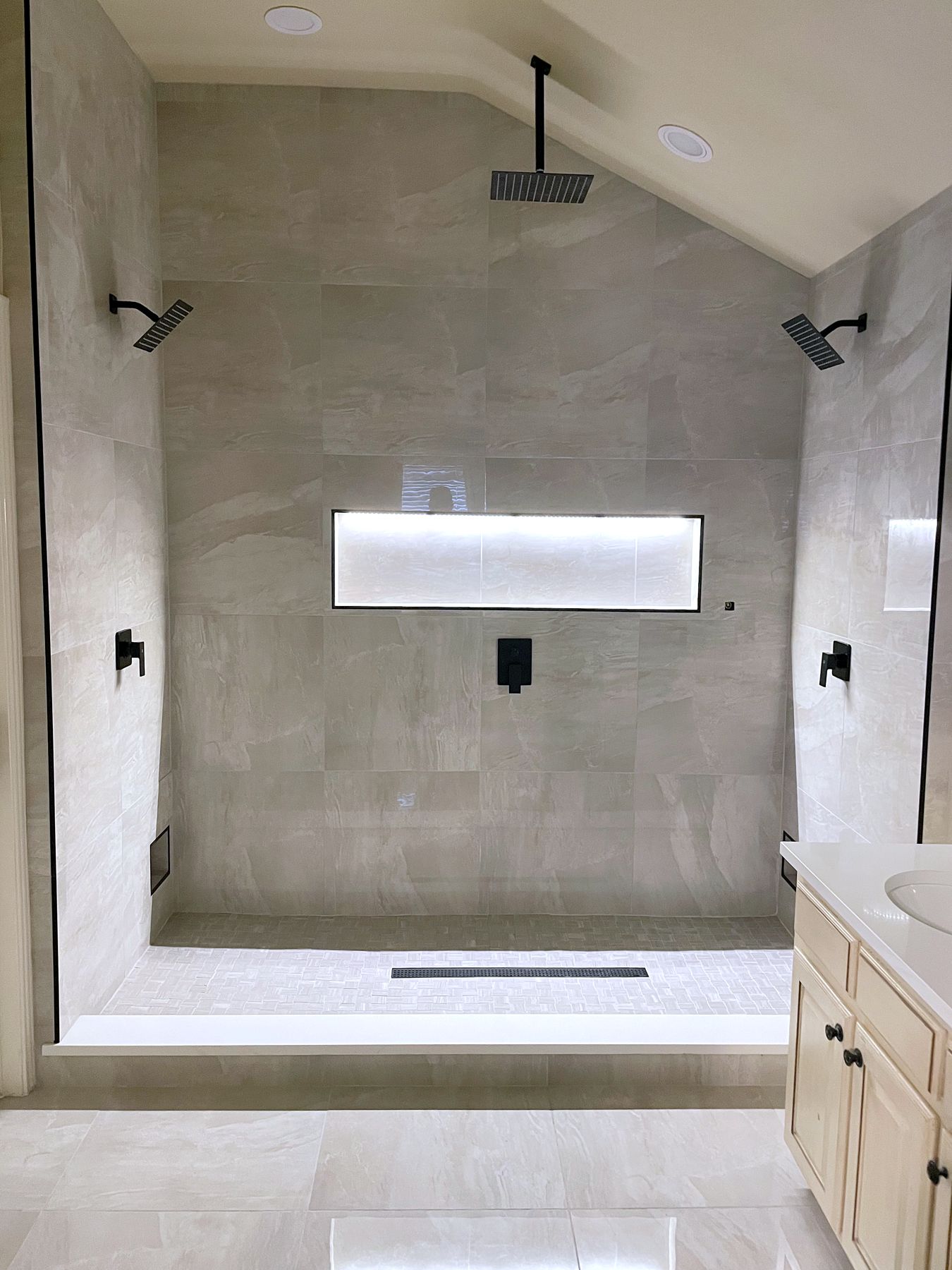 Walk-in shower with matte black fixtures, a ceiling rain head, two wall heads and a lit niche
