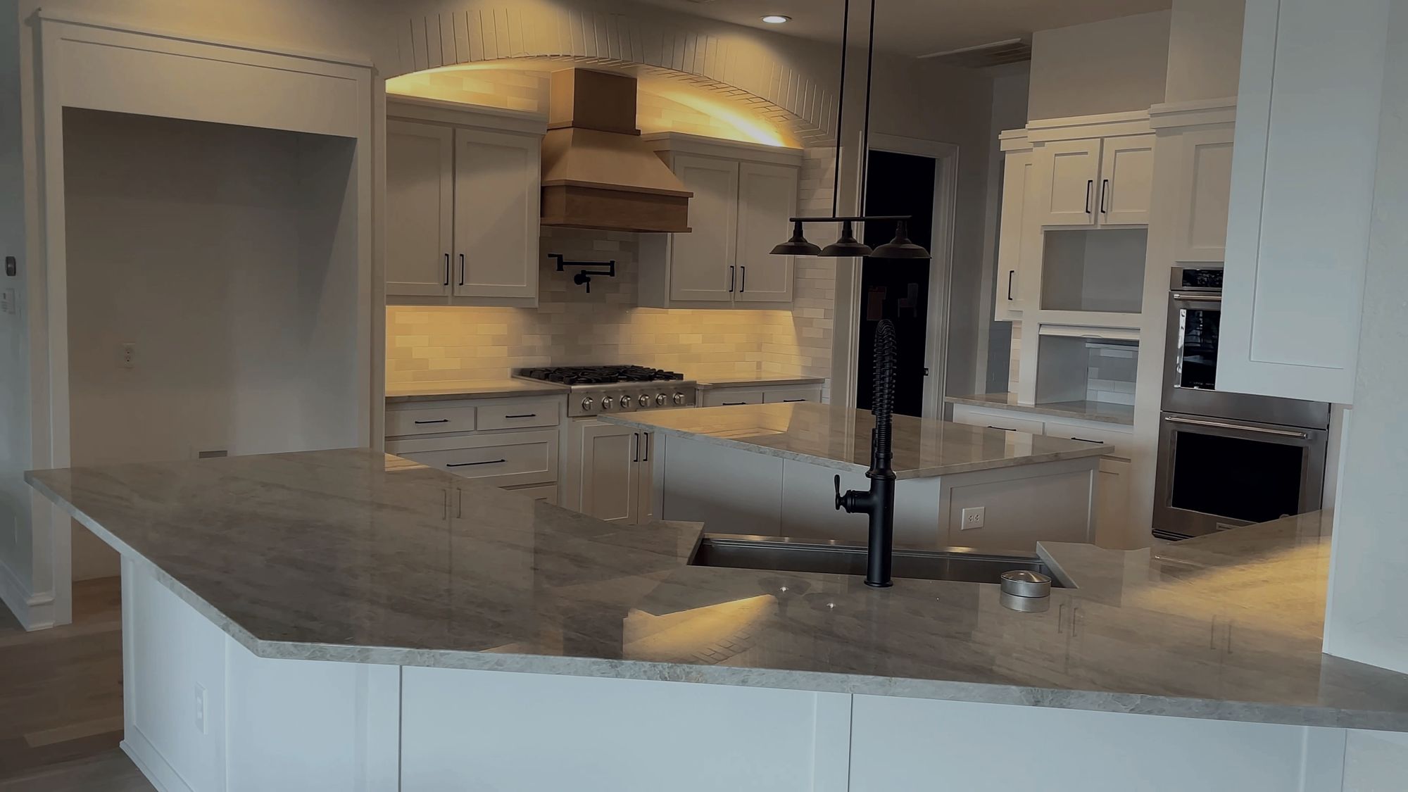 Kitchen with a cove-lit brick arch over a white oak hood, pot filler, two stone-topped islands and double wall ovens