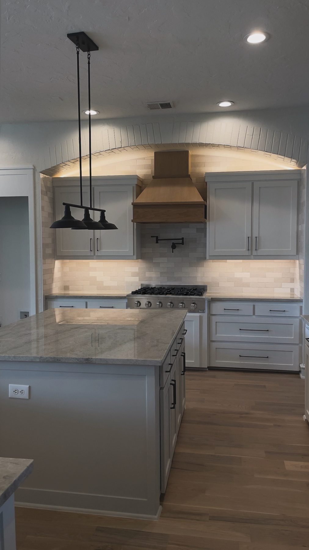 Kitchen with a cove-lit brick arch, white oak range hood, stainless gas rangetop and stone island